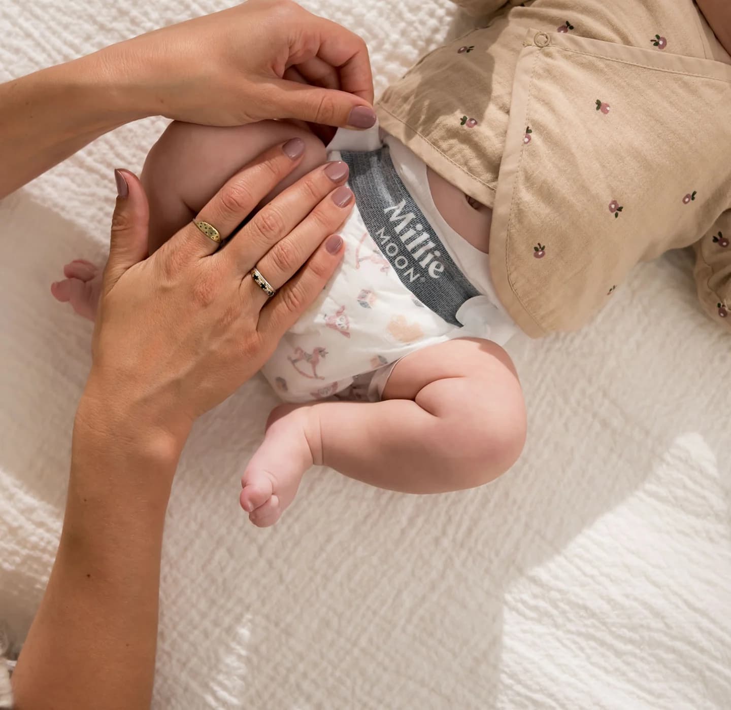 Parent fastening the waistband tab of a Millie Moon Luxury Tape Diaper on a baby lying on a bed