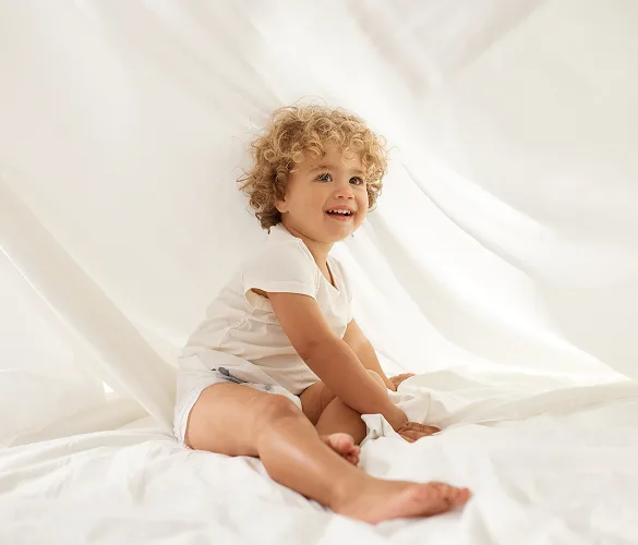 Smiling toddler in a Millie Moon diaper sitting among soft white sheets