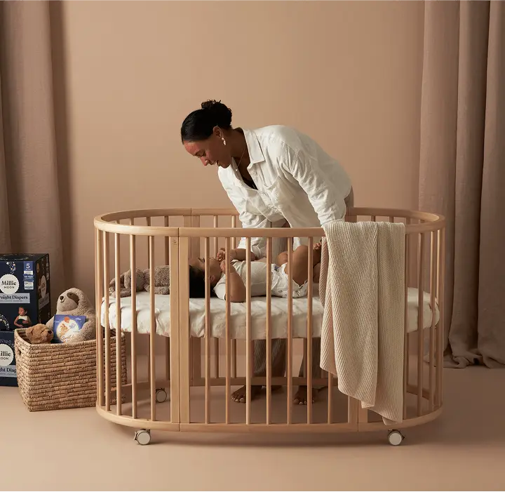 Parent settling a baby into a crib beside a pack of Millie Moon Overnight Tape Diapers
