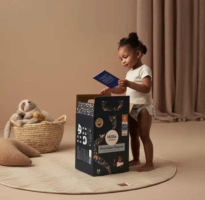 Toddler reading a Millie Moon storybook beside a box of Millie Moon Overnight Tape Diapers
