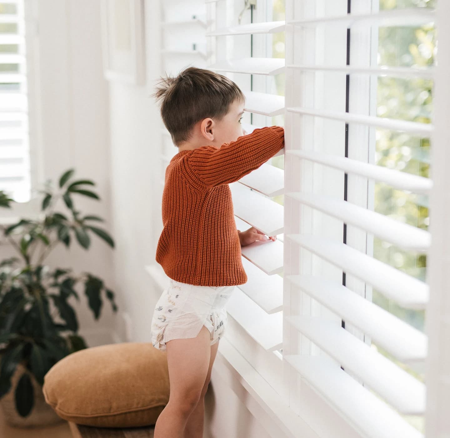 Toddler in a knit jumper and a Millie Moon tape diaper peering through white window shutters